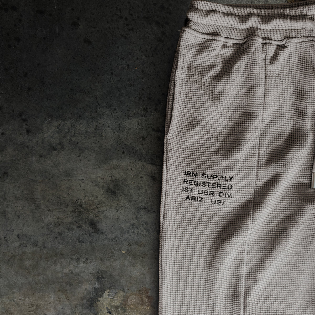 A close-up of light beige textured sweatpants laid flat on a dark concrete surface, featuring black printed text on the thigh.