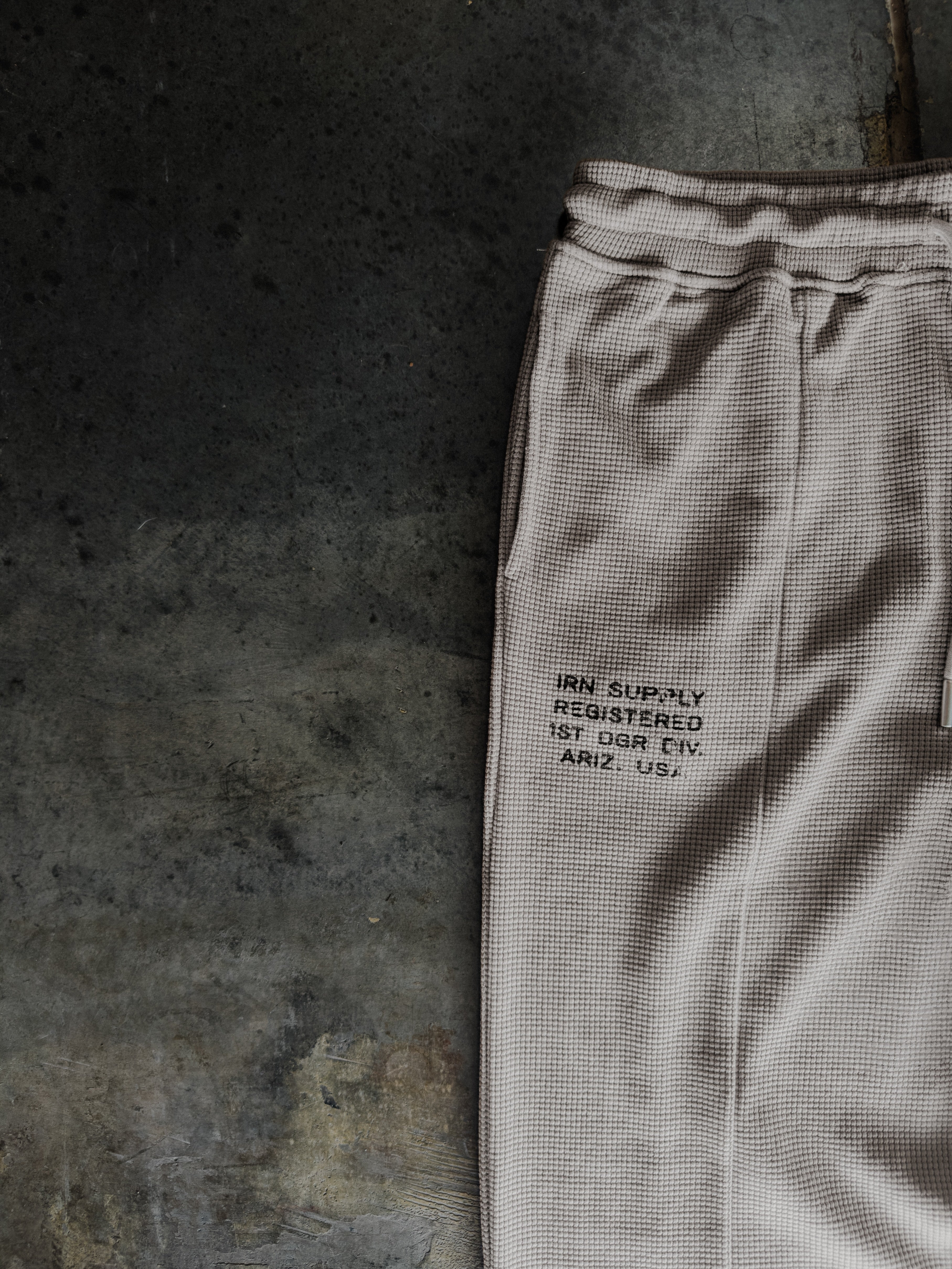 A close-up of light beige textured sweatpants laid flat on a dark concrete surface, featuring black printed text on the thigh.