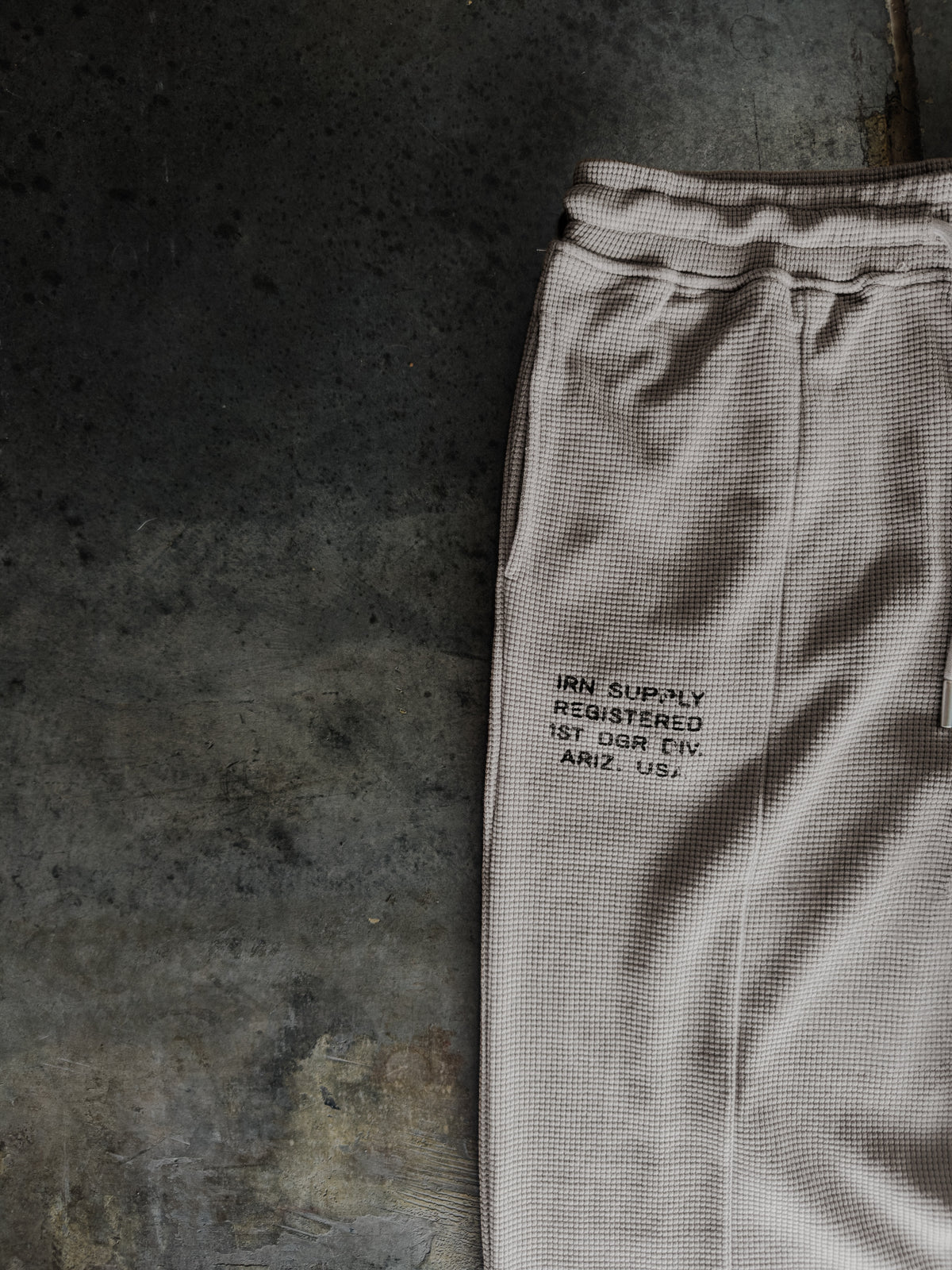 A close-up of light beige textured sweatpants laid flat on a dark concrete surface, featuring black printed text on the thigh.